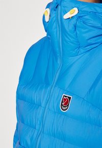 Blauwe pufferjack met een rits aan de voorkant, hoge kraag en een logo patch met een rode vos. Zacht, textuurstof met horizontale naden.