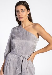 Asymmetrische top met één schouder van textuurstof in lichtgrijs, met een ceintuurlijfje en een speelse pofmouw. Zilveren studsieraden maken de look compleet.