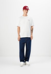 Hvid bomulds t-shirt med et lille, rødt logo, parret med blå denim jeans. En rød kasket og hvide sneakers fuldender outfittet.