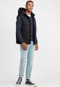 Schwarze Daunenweste mit Kapuze, Reißverschluss, getragen über einem marineblauen Pullover, kombiniert mit hellblauen Jeans und marineblauen Sneakers. Einfaches Design, keine sichtbaren Muster.