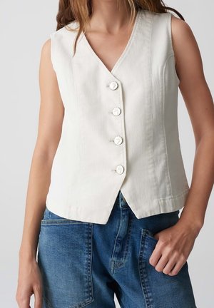 Femme portant un gilet blanc sans manches boutonné, associé à un jean denim bleu, main dans la poche, sur un fond uni.