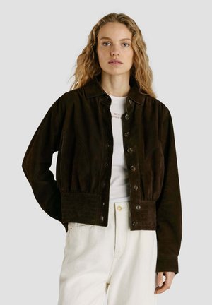 Donna con capelli mossi che indossa una giacca marrone scuro con bottoni sopra una t-shirt bianca e pantaloni a vita alta di colore chiaro su uno sfondo semplice.