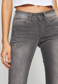Jeans desgastados grises con cinco bolsillos, cierre de botón y detalles de costura en contraste. La tela parece tener textura con acentos desgastados.