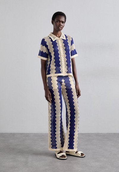 Crochet knit co-ord set in cream with blue patterns, featuring a collared shirt and wide-leg pants, worn with flat sandals.