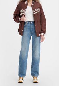 Veste bomber en satin marron avec des lettres blanches, associée à un jean taille haute bleu clair et des baskets à motifs. Col et poignets rayés.