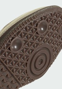 Suola di scarpa beige con superficie in gomma testurizzata caratterizzata da motivi triangolari e accenti circolari. Il bordo presenta un design rialzato e dettagliato.