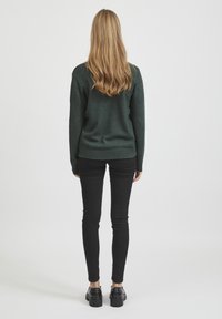 Maglione lavorato a maglia verde scuro con orlo a costine, indossato sopra jeans neri attillati e scarpe nere, visto da dietro.