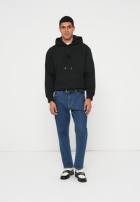 Sweatshirt preta com cordões e bolso frontal, combinada com jeans azuis e sapatos pretos e brancos. Design simples e forma ajustada.