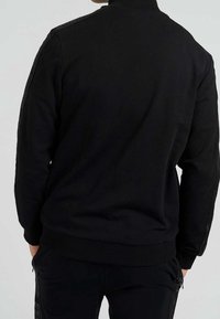 Sweat-shirt noir avec un col montant, des poignets et un ourlet côtelés. Présente des panneaux latéraux texturés, offrant un design en couches.