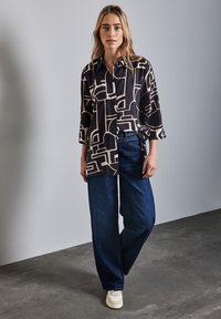 Camicia nera con bottoni e un motivo geometrico crema, abbinata a jeans larghi di un blu scuro e scarpe da ginnastica chiare su uno sfondo grigio.