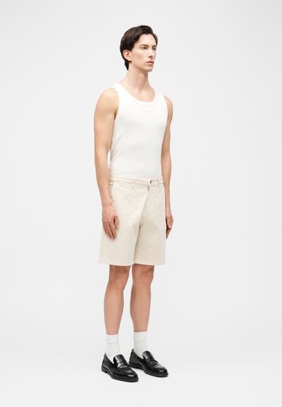 Άνδρας που φοράει λευκή αμάνικη φανέλα, μπεζ σορτς μήκους μέχρι το γόνατο, λευκές κάλτσες και μαύρα δερμάτινα μοκασίνια, στέκεται μπροστά σε απλό φόντο.