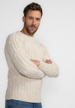Cream cable-knit sweater with a round neck, textured pattern, and ribbed cuffs and hem. Worn with blue jeans, arms crossed.