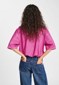 Ljust rosa överstor skjorta med korta ärmar, tillverkad av ett mjukt tyg. Kombineras med löst sittande blå denimjeans med bakfickor.