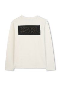 Chemise blanche à manches longues avec un panneau rectangulaire noir au dos portant le mot "KARL" en couture marron. Texture de tissu douce.