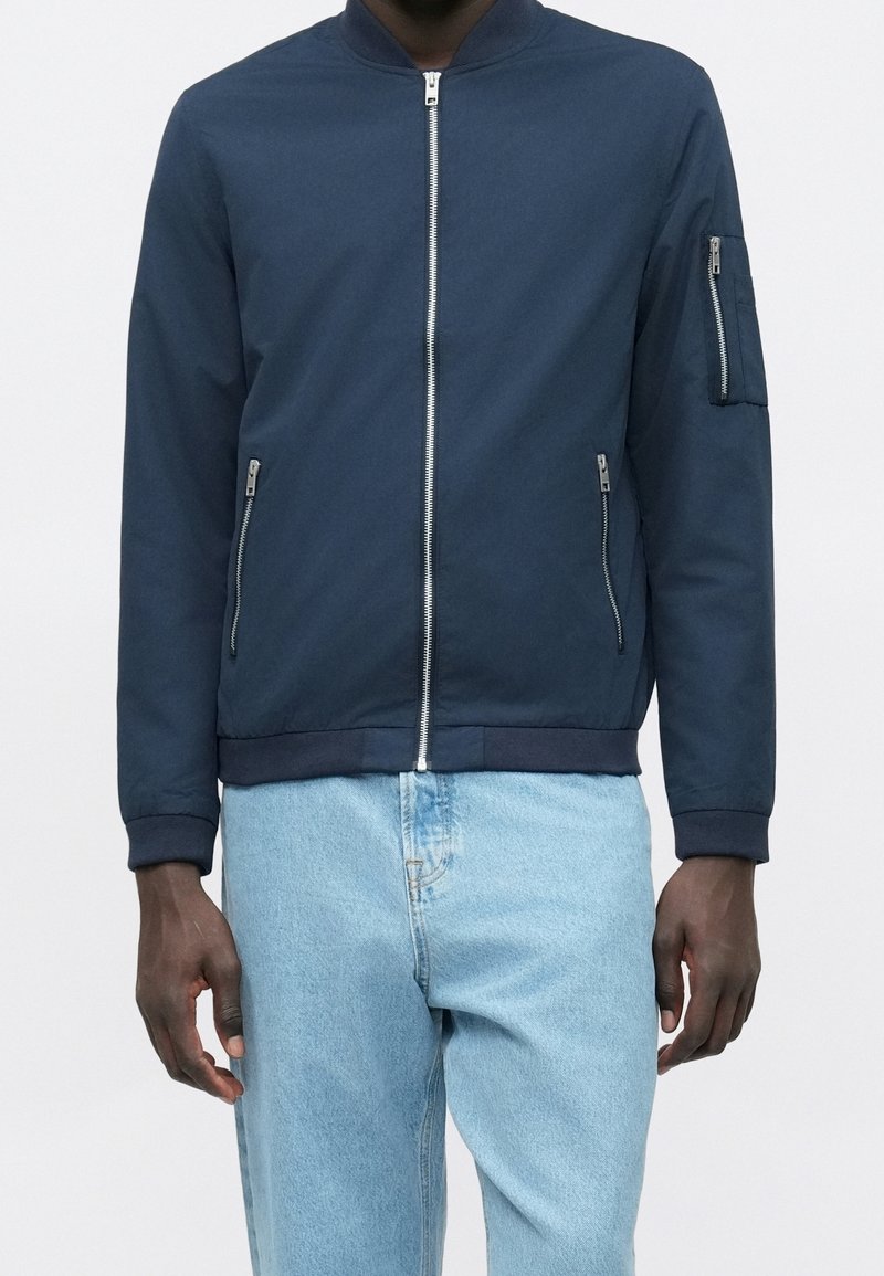 Homme portant une veste bomber bleu marine avec des poches zippées devant et sur les manches, associée à un jean bleu clair sur un fond uni.