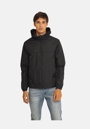 Homme portant une veste noire à capuche zippée avec des poignets élastiques et un jean bleu clair, debout devant un fond blanc uni.