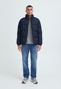Mørkeblå pufferjakke med høj krave, parret med en grå sweater og blå jeans. Tekstureret stof og polstret design er synligt.