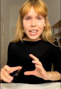 Junge Frau mit blonden Haaren und schwarzem Top, die spricht und mit den Händen gestikuliert, sitzt drinnen an einem Schreibtisch und Regalen.