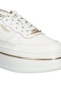 Baskets blanches en cuir avec une semelle plateforme blanche épaisse, bande accent dorée et détail de logo sur la languette. Les lacets sont plats et blancs.