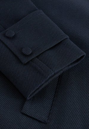 Dunkelblauer, strukturiertes Gewebe mit feinem Streifenmuster. Verfügt über einen Umschlag mit zwei Knöpfen und einem spitzen Klappendetail.