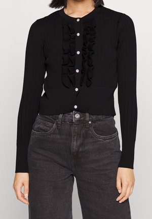Cardigan noir à côtes avec une fermeture à boutons à l'avant, un détail volanté au centre et des manches longues. Porté avec un jean gris foncé.