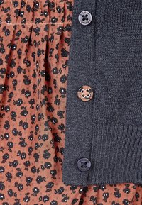 Tmavě modrý pletený kardigan vrstvený přes rezavě hnědé tričko s květinovým vzorem a černými květy. Má dva knoflíky, jeden s označením značky.