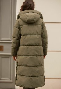 Manteau matelassé vert olive, longueur genoux avec capuche, présentant des sections matelassées horizontales, des fentes sur les côtés et une texture lisse et mate.