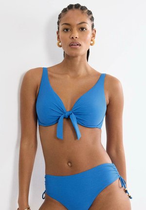 Frau mit geflochtenem Haar, die einen blauen zweiteiligen Badeanzug mit vorderem Knotenoberteil und gerafften Seitenschnürungen vor weißem Hintergrund trägt.