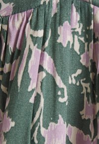 Tecido verde com padrões florais em roxo e branco, apresentando pregas que criam textura e profundidade. Material suave e tecido.