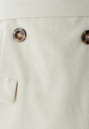 Šviesiai kreminės spalvos audinys su matoma tekstūra, kuriame yra du dideli gintariniai mygtukai ir plokščia kišenė kairėje pusėje.