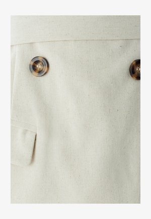 Šviesiai kreminės spalvos audinys su matoma tekstūra, kuriame yra du dideli gintariniai mygtukai ir plokščia kišenė kairėje pusėje.