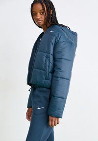 Zkrátená tmavomodrá péřová bunda s kapucňou, s predným zipsom a elastickými manžetami. Ľahký materiál s hladkou textúrou. Viditeľné logo Nike.
