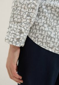 Leichtes Shirt mit einem grau-weißen abstrakten Muster, mit Knopfmanschetten und einem abgerundeten Saum, kombiniert mit dunklen Hosen.