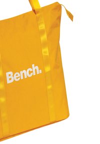 Gele draagtas van een gladde, duurzame stof. Voorzien van brede, bijpassende schouderbanden en opvallende witte tekst met "Bench." aan de voorkant.