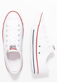 Bílé plátěné tenisky s červenými a modrými pruhy podél podešve. Mají kovové průvlaky a texturovaný gumový špičku. Viditelné logo Converse.
