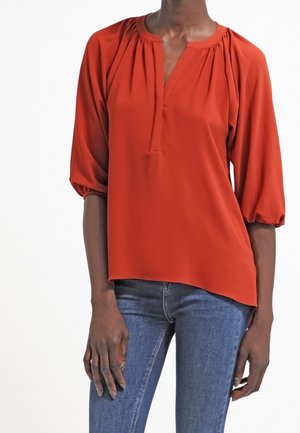 Kvinde iført en løs, rust-orange bluse med 3/4 puffede ærmer og blå jeans, stående mod en hvid baggrund.