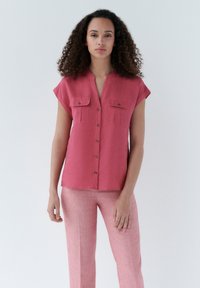 Lichtroze blouse met korte mouwen en twee borstzakken, gecombineerd met lichte roze broek, beiden in een zachte stof met een subtiele textuur.