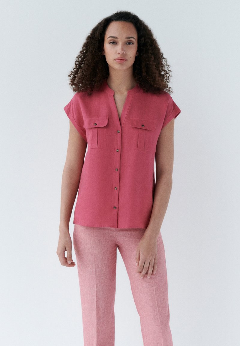 Lichtroze blouse met korte mouwen en twee borstzakken, gecombineerd met lichte roze broek, beiden in een zachte stof met een subtiele textuur.