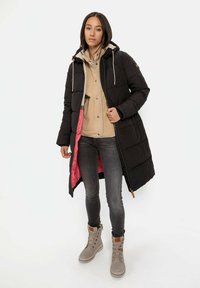Zwarte pufferjas met een middellange ontwerp, voorzien van een roze voering, een capuchon met trekkoord en zijzakken, gedragen over een beige hoodie en grijze spijkerbroek.