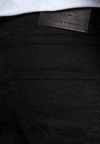 Vaqueros de mezclilla negra con una textura suave, que cuentan con un bolsillo trasero y una etiqueta de cuero cosida en la cinturilla con texto en relieve.