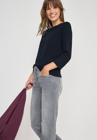 Maglietta a maniche lunghe blu navy con scollatura rotonda, abbinata a jeans in denim grigio chiaro. Tiene un indumento bordeaux, ambientato su uno sfondo chiaro.