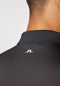 Polo nero con colletto a coste. Presenta un piccolo logo bianco sul retro. Texture liscia e design aderente.