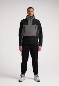 Giacca impermeabile da uomo con pannelli grigi e neri, tasche con zip e cappuccio regolabile. Abbinata a pantaloni cargo neri e sneakers grigie.