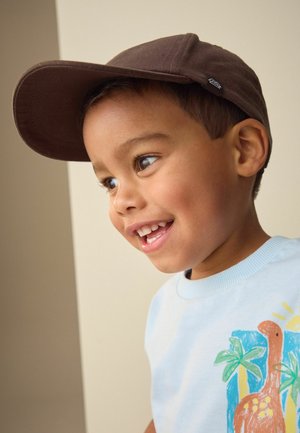 Ung dreng smiler og kigger til højre, iført en brun kasket og en lyseblå skjorte med et farverigt dinosaur- og palmetræsdesign.