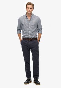 Giovane uomo in piedi con le mani nelle tasche che indossa una camicia a quadri con bottoni, pantaloni scuri, cintura marrone, orologio e scarpe nere su sfondo bianco.