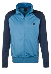 Bunda na zip v modré a námořnické barvě. Má stojáček, ribbed manžety a nášivku s logem na levé hrudi. Hladký povrch.