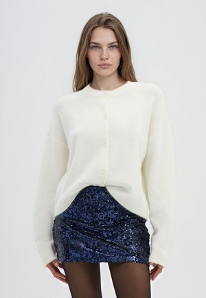 Giovane donna che indossa un maglione bianco con bottoni, una mini gonna di paillettes blu scuro e calze nere, in piedi contro uno sfondo chiaro e uniforme.