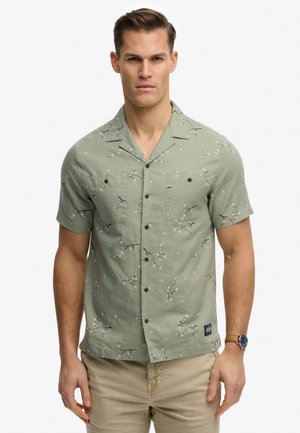 Hombre vistiendo una camisa de manga corta de color verde claro con estampado floral y pantalones beige, mirando hacia adelante con expresión neutral.