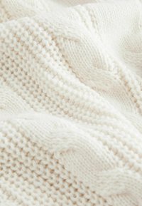 Weiße Strickdecke mit weichem Textur, die abwechselnde Muster aus engen und lockeren Maschen aufweist, was ein gemütliches, geschichtetes Aussehen erzeugt.