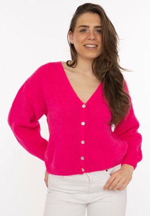 Junge Frau lächelt, trägt eine leuchtend pinke, geknöpfte Strickjacke und weiße Hose, mit langen, welligen braunen Haaren über der Schulter.